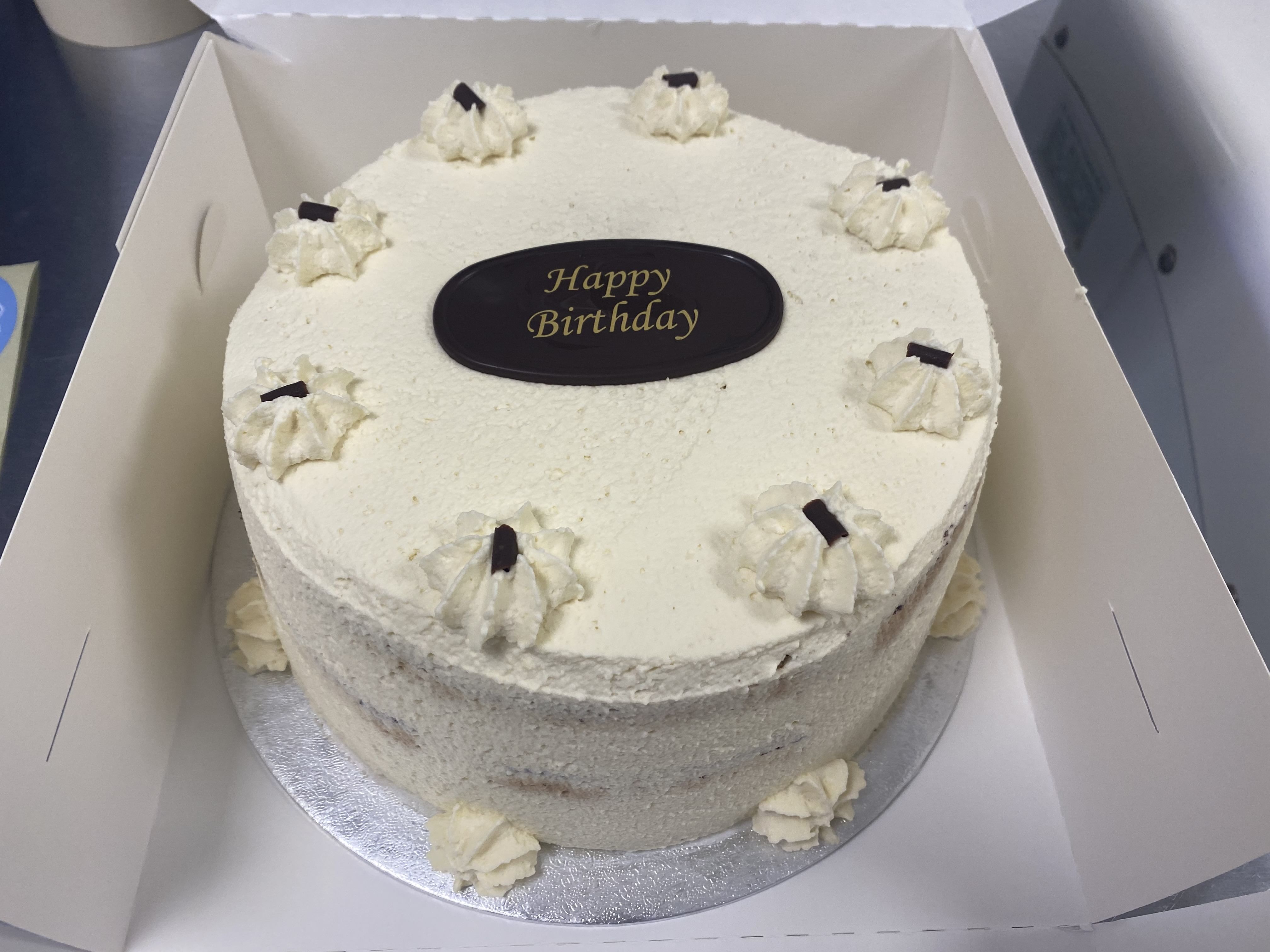 A round, white-frosted Black Forest gateaux rests inside an open box. It is decorated with dollops of whipped cream around the top and tiny chocolate shavings on each dollop. A chocolate plaque in the center reads 'Happy Birthday.' The cake, infused with subtle cherry liqueur, sits on a silver board.